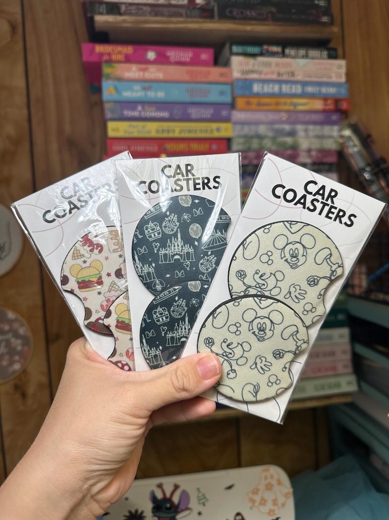 May include: Three sets of car coasters featuring different designs. The first set has a black and white design with a castle and other Disney park icons. The second set has a black and white design with Mickey Mouse and his signature gloves. The third set has a colorful design with a hamburger, a Mickey Mouse head, and a red and white striped design.