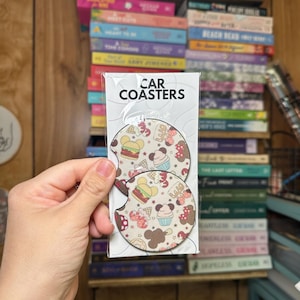 May include: Two round car coasters with a white background and a colorful pattern of cartoon food and Mickey Mouse ears. The text "CAR COASTERS" is printed on the packaging.