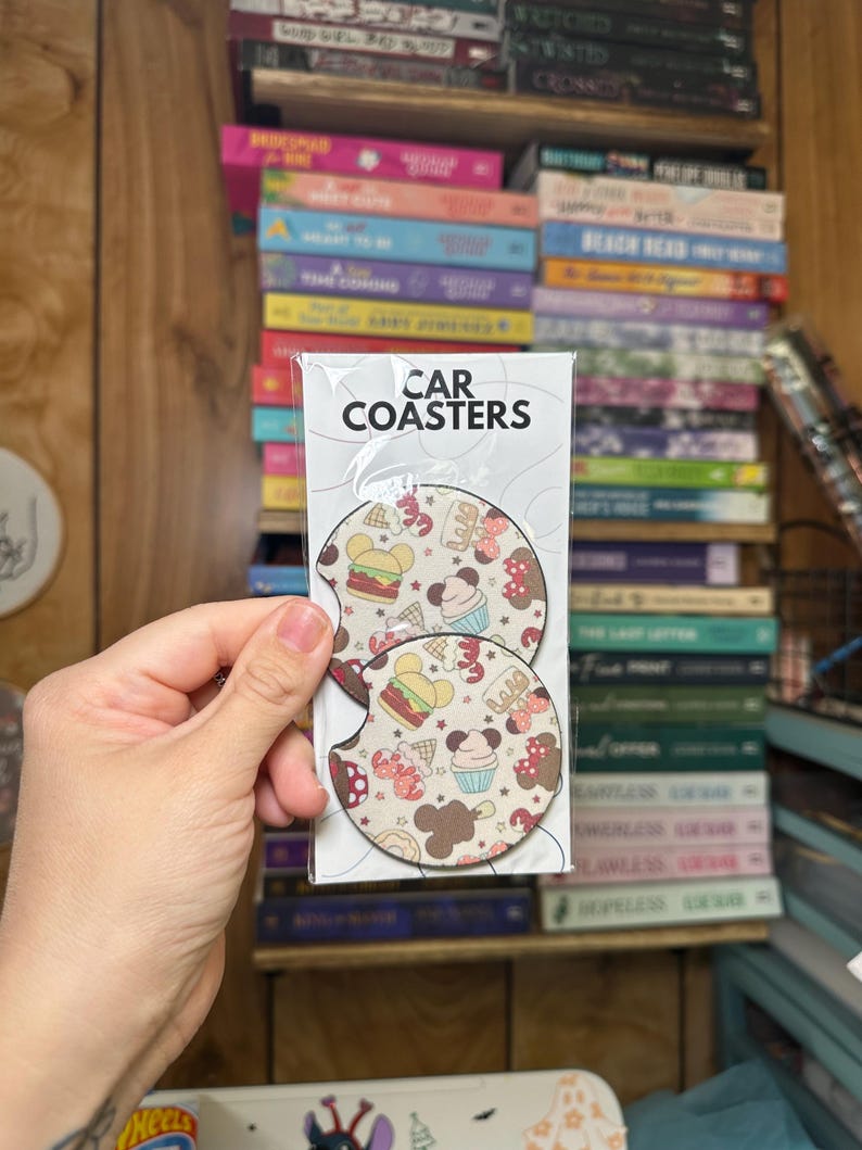 May include: Two round car coasters with a white background and a colorful pattern of cartoon food and Mickey Mouse ears. The text "CAR COASTERS" is printed on the packaging.