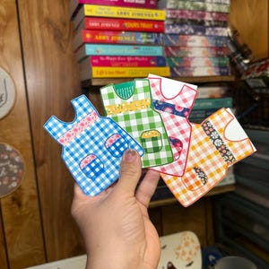 May include: Four paper doll dresses with different patterns. The dresses are blue and white gingham, green and white gingham, red and white gingham, and orange and white gingham. Each dress has a floral pattern around the waist.
