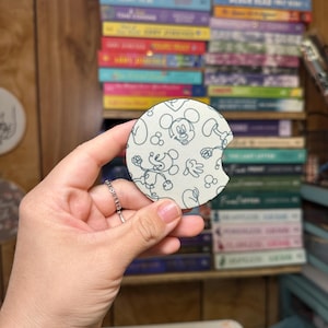 May include: A white and blue round coaster with a black and white outline of Mickey Mouse. The coaster has a white background with black and white outlines of Mickey Mouse's head and body. The coaster is made of a soft material and has a slightly textured surface.
