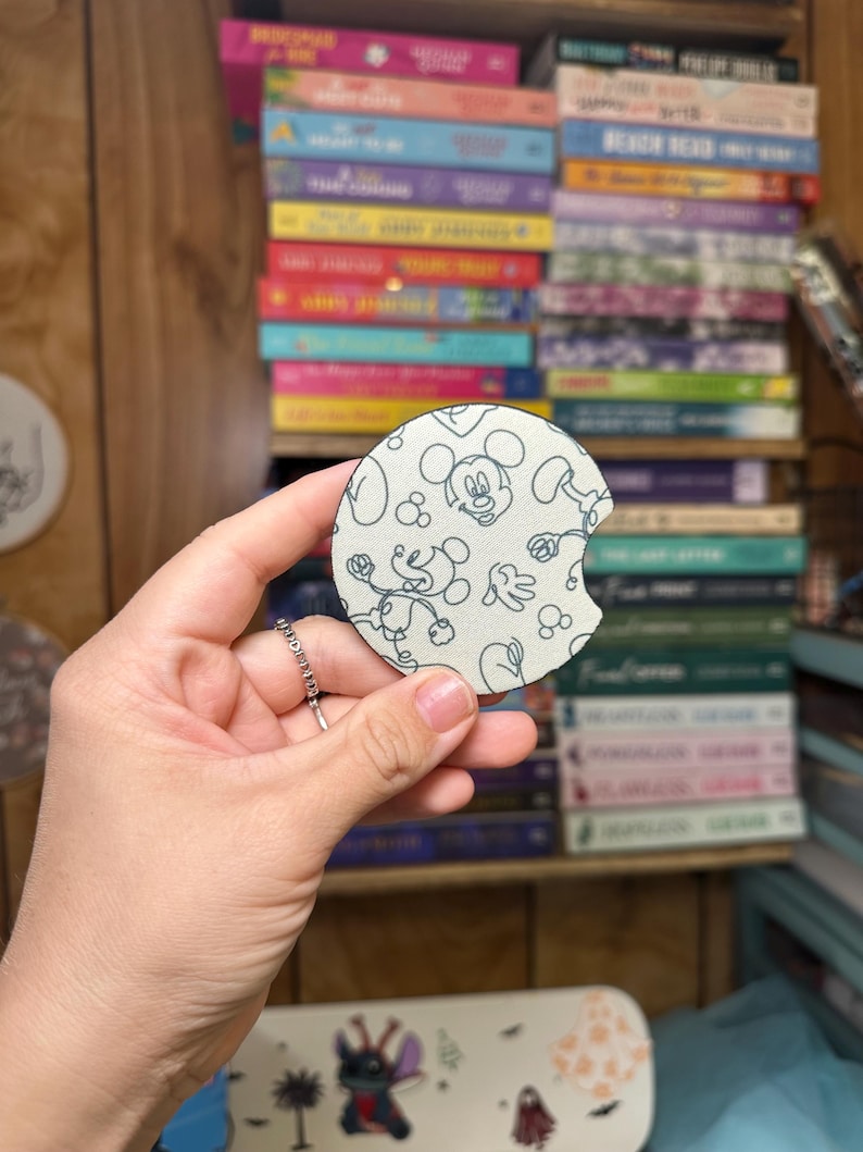 May include: A white and blue round coaster with a black and white outline of Mickey Mouse. The coaster has a white background with black and white outlines of Mickey Mouse's head and body. The coaster is made of a soft material and has a slightly textured surface.