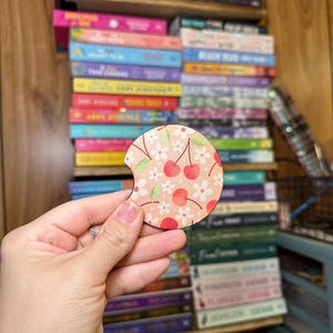 May include: A round, wooden coaster with a cherry and floral design. The coaster has a cutout for a drink. The coaster is pink, white, and green.
