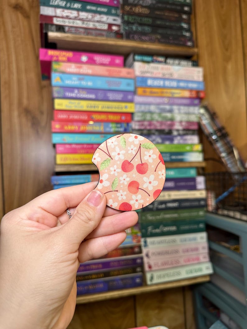 May include: A round, wooden coaster with a cherry and floral design. The coaster has a cutout for a drink. The coaster is pink, white, and green.