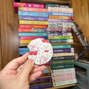 May include: A round white coaster with a cherry and bow print. The coaster has a pink and white pattern with red cherries and pink bows.