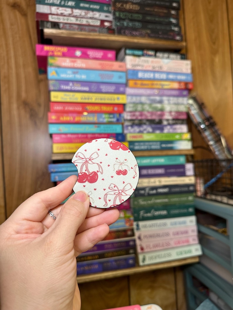 May include: A round white coaster with a cherry and bow print. The coaster has a pink and white pattern with red cherries and pink bows.
