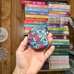 May include: A round, blue and gray patterned coaster with a red and blue Spiderman design. The coaster features multiple images of Spiderman in different poses.