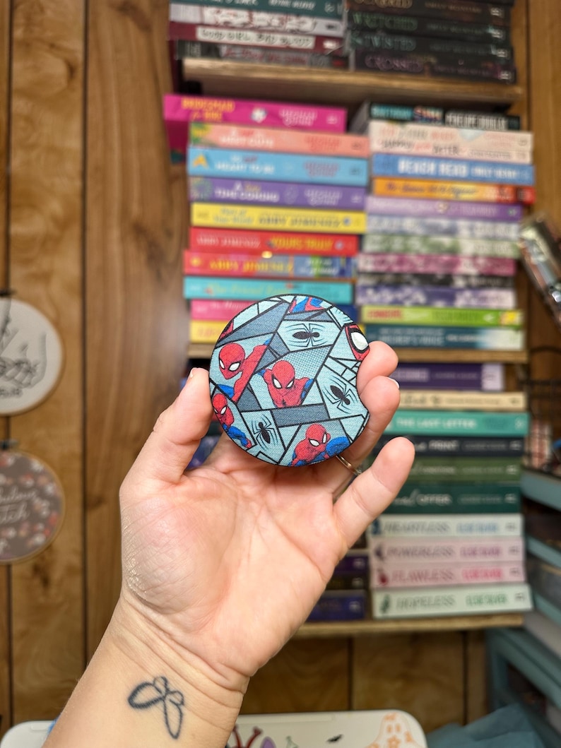 May include: A round, blue and gray patterned coaster with a red and blue Spiderman design. The coaster features multiple images of Spiderman in different poses.