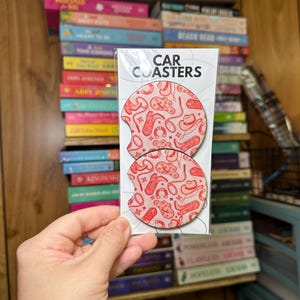 May include: Two round car coasters with a pink and red cowboy themed design. The coasters are packaged in a clear plastic sleeve with the text "CAR COASTERS" printed on the front.