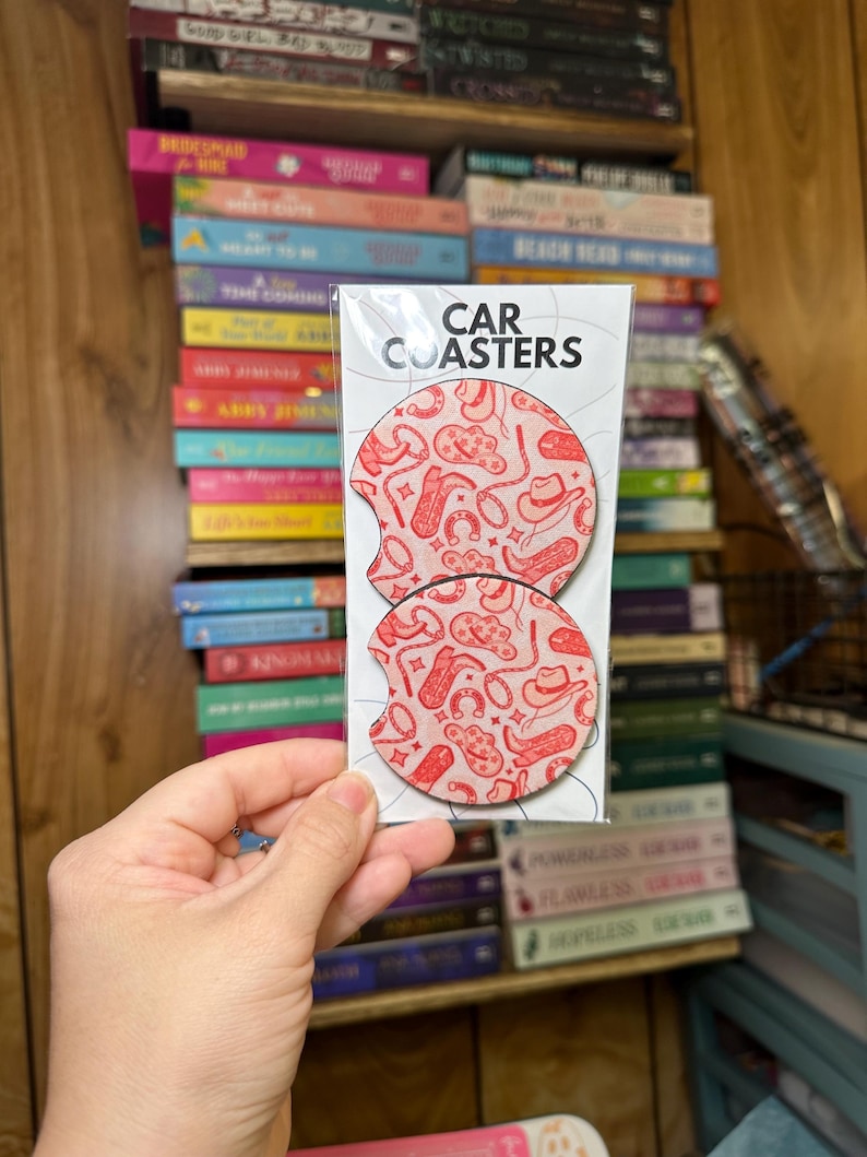May include: Two round car coasters with a pink and red cowboy themed design. The coasters are packaged in a clear plastic sleeve with the text "CAR COASTERS" printed on the front.