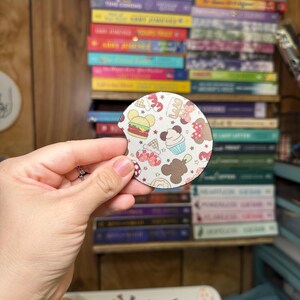 May include: A round, white coaster with a colorful pattern of cartoon food and drinks. The coaster features a Mickey Mouse head shape with a red bow and a white polka dot pattern. The food and drinks include a hamburger, a cupcake, a Mickey Mouse ice cream cone, a donut, and a Mickey Mouse pretzel.