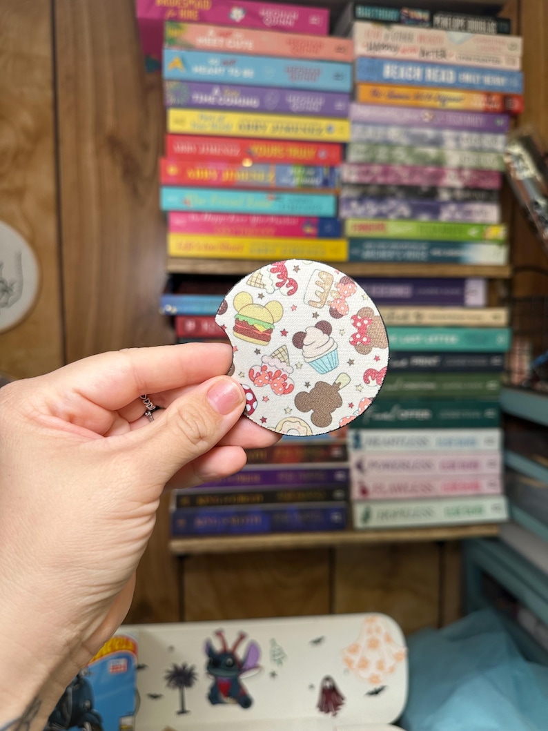 May include: A round, white coaster with a colorful pattern of cartoon food and drinks. The coaster features a Mickey Mouse head shape with a red bow and a white polka dot pattern. The food and drinks include a hamburger, a cupcake, a Mickey Mouse ice cream cone, a donut, and a Mickey Mouse pretzel.