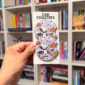 May include: Two circular car coasters in a clear plastic sleeve with the text CAR COASTERS at the top. The coasters feature a light purple background with a repeating pattern of Snoopy dressed as a vampire, Woodstock in a witch hat, ghosts, and orange jack-o'-lanterns.