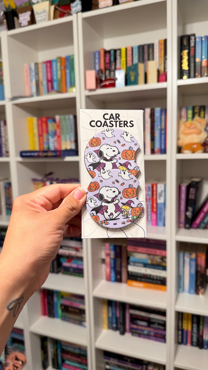 May include: Two circular car coasters in a clear plastic sleeve with the text CAR COASTERS at the top. The coasters feature a light purple background with a repeating pattern of Snoopy dressed as a vampire, Woodstock in a witch hat, ghosts, and orange jack-o'-lanterns.