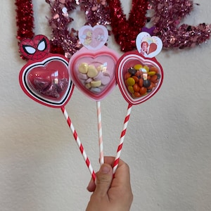 May include: Three heart-shaped candy holders on sticks, filled with sweets. The holders are red and white, with character stickers. The word "LOVE" is spelled out in red tinsel in the background.