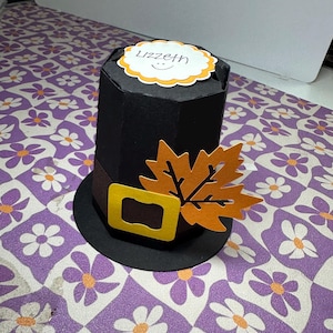 Könnte beinhalten: Ein schwarzer Papier-Pilgerhut mit einer gelben Schnalle, einem braunen Gürtel und einem orangefarbenen Papierblatt. Der Hut hat einen weißen Kreis oben mit dem Namen "Lizzeth" in Schwarz.