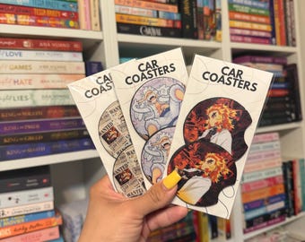 May include: Three car coasters are held in a hand, each with a unique design. One features a character with orange hair and a red and black background. Another has a white and blue character. The third has a wanted poster design.