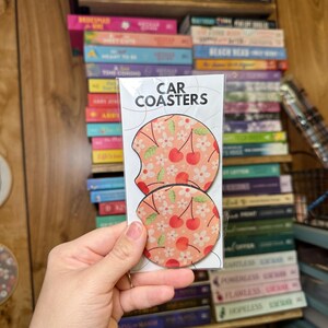 May include: Two round car coasters with a cherry and floral print. The coasters are pink with white flowers and red cherries. The text "CAR COASTERS" is printed on the packaging.