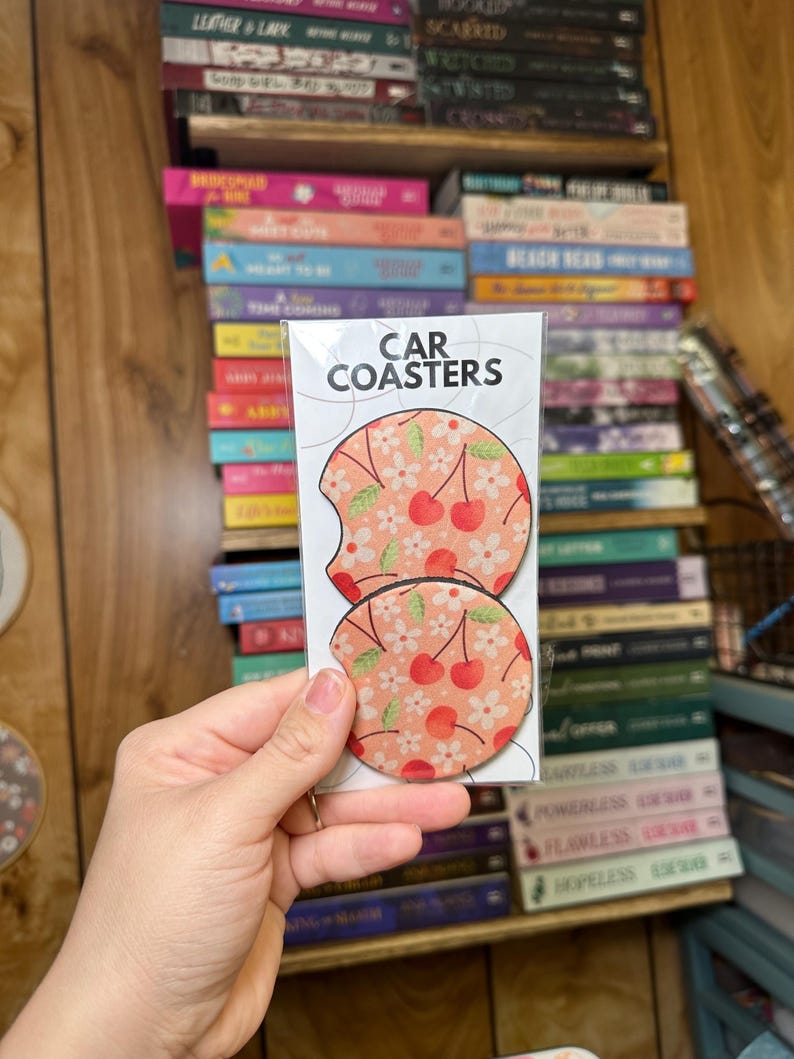 May include: Two round car coasters with a cherry and floral print. The coasters are pink with white flowers and red cherries. The text "CAR COASTERS" is printed on the packaging.