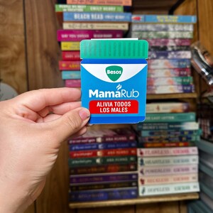 May include: A blue and white sticker with the text "MamaRub" and "Alivia Todos Los Males" in a blue box. The sticker is shaped like a bottle with a green lid and the text "Besos" in a white box.