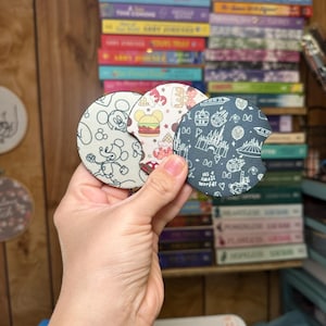 May include: Three round, fabric coasters with different Disney designs. The first coaster features Mickey Mouse and Minnie Mouse with a Mickey Mouse ice cream bar. The second coaster features a Mickey Mouse shaped hamburger and a Mickey Mouse ice cream bar. The third coaster features a black and white design of Cinderella's castle, a carousel, and a fireworks display. The text "It's a small world" is printed on the coaster.