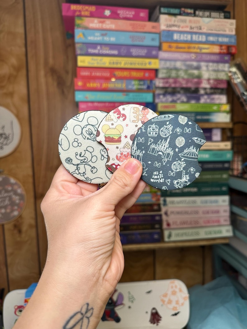 May include: Three round, fabric coasters with different Disney designs. The first coaster features Mickey Mouse and Minnie Mouse with a Mickey Mouse ice cream bar. The second coaster features a Mickey Mouse shaped hamburger and a Mickey Mouse ice cream bar. The third coaster features a black and white design of Cinderella's castle, a carousel, and a fireworks display. The text "It's a small world" is printed on the coaster.