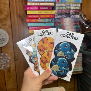 May include: Three sets of car coasters featuring cartoon characters. The coasters are made of wood and have a glossy finish. The first set features a blue and green character, the second set features an orange character, and the third set features a blue character. Each set has the words "Car Coasters" printed on the packaging.