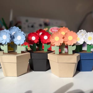 Könnte beinhalten: Eine Sammlung von Papierblumenarrangements in verschiedenen Farben, darunter Blau, Rot, Orange und Weiß. Jede Blume befindet sich in einem geometrischen Papiertopf. Die Töpfe sind beige, schwarz und blau.