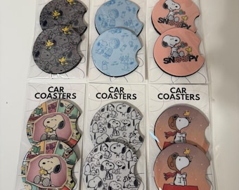 May include: Six sets of two circular car coasters in clear plastic packaging, each labeled CAR COASTERS. The designs feature Snoopy and Woodstock in various patterns, including gray, blue, peach, comic strip, black and white, and aviator themes.