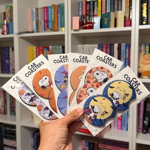 May include: A collection of circular car coasters packaged in clear plastic sleeves, each featuring Snoopy and Woodstock in various seasonal and holiday-themed illustrations, with the text CAR COASTERS printed on the white backing cards.