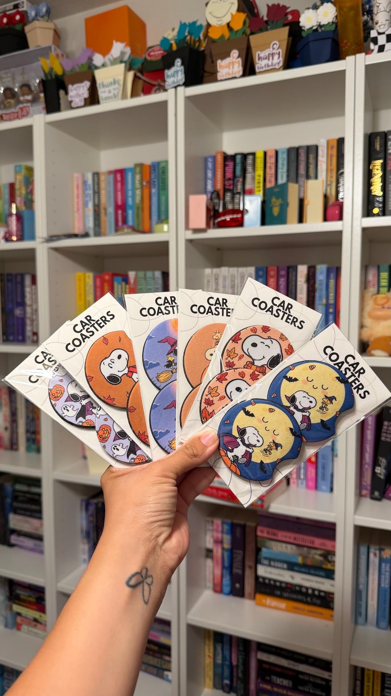 May include: A collection of circular car coasters packaged in clear plastic sleeves, each featuring Snoopy and Woodstock in various seasonal and holiday-themed illustrations, with the text CAR COASTERS printed on the white backing cards.
