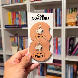 May include: Two round, peach-colored car coasters featuring a graphic of Snoopy and Woodstock sitting inside a carved jack-o'-lantern pumpkin. The coasters are displayed on a white backing card with the text CAR COASTERS printed at the top.