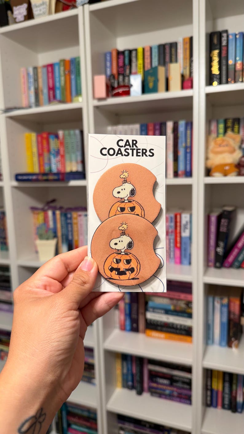 May include: Two round, peach-colored car coasters featuring a graphic of Snoopy and Woodstock sitting inside a carved jack-o'-lantern pumpkin. The coasters are displayed on a white backing card with the text CAR COASTERS printed at the top.