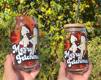 May include: Two clear glass tumblers with a red, white, and green design. The tumblers have the text "Merry Fetchmas" on them. One tumbler has a straw in it.