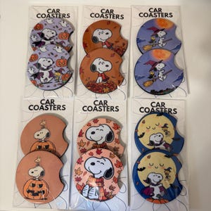 May include: Six sets of two circular car coasters in clear plastic packaging, each labeled CAR COASTERS. The coasters feature Snoopy and Woodstock in various seasonal designs, including Halloween pumpkins, autumn leaves, and witch-themed imagery.