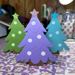 Puede incluir: Tres árboles de Navidad de papel, uno morado, uno verde y uno azul, con lunares blancos y estrellas brillantes en la parte superior.