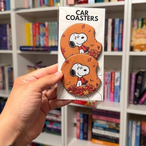 May include: Two round car coasters in a clear plastic sleeve with a white header card labeled CAR COASTERS. Each coaster features a cartoon illustration of Snoopy wearing a red scarf, sitting in a pile of orange and red autumn leaves.