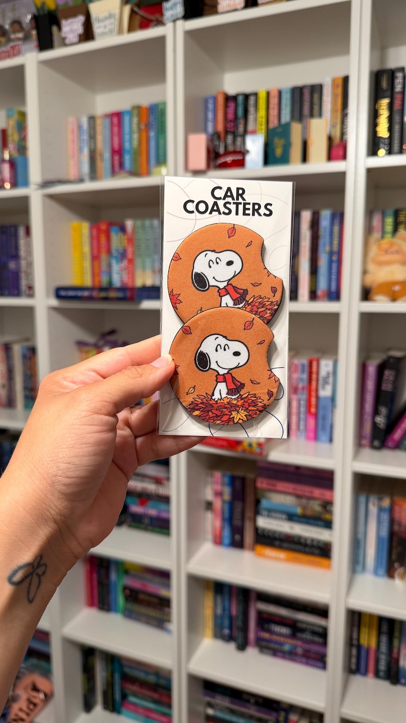 May include: Two round car coasters in a clear plastic sleeve with a white header card labeled CAR COASTERS. Each coaster features a cartoon illustration of Snoopy wearing a red scarf, sitting in a pile of orange and red autumn leaves.