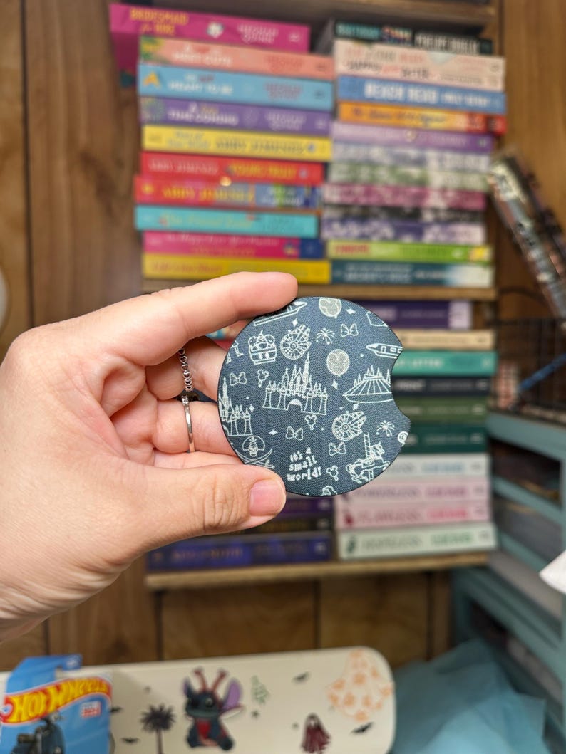May include: A black and white car coaster with a design featuring various Disney attractions, including Cinderella's Castle, Space Mountain, and the 'it's a small world' ride. The coaster has a black background and white outlines of the attractions.