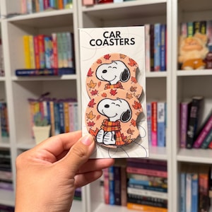 May include: Two round car coasters on a white display card with the text CAR COASTERS. Each coaster features a cartoon dog with black ears wearing a yellow and orange striped scarf, surrounded by a pattern of orange, red, and yellow autumn leaves.