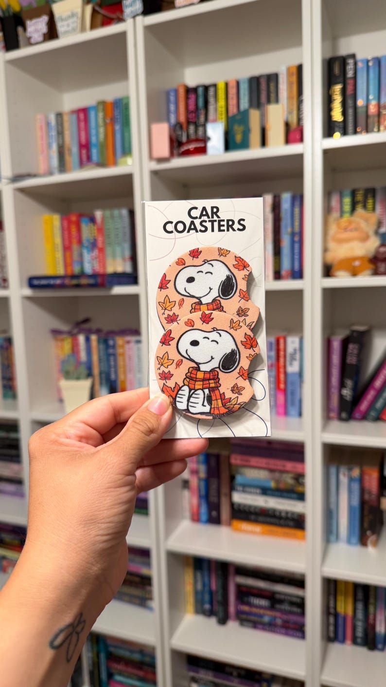 May include: Two round car coasters on a white display card with the text CAR COASTERS. Each coaster features a cartoon dog with black ears wearing a yellow and orange striped scarf, surrounded by a pattern of orange, red, and yellow autumn leaves.