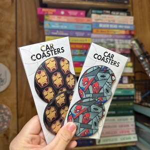 May include: Two sets of car coasters featuring popular superhero characters. One set features Iron Man's mask in a repeating pattern on a black background. The other set features Spider-Man in a repeating pattern on a blue and gray background. The coasters are packaged in clear plastic.