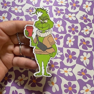May include: A green cartoon character with a white collar and a brown satchel, holding a red cup of coffee. The character is standing on a purple and white floral background.