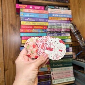 May include: Two round, pink and white coasters with a cherry and bow design. The coasters are made of wood and have a glossy finish.
