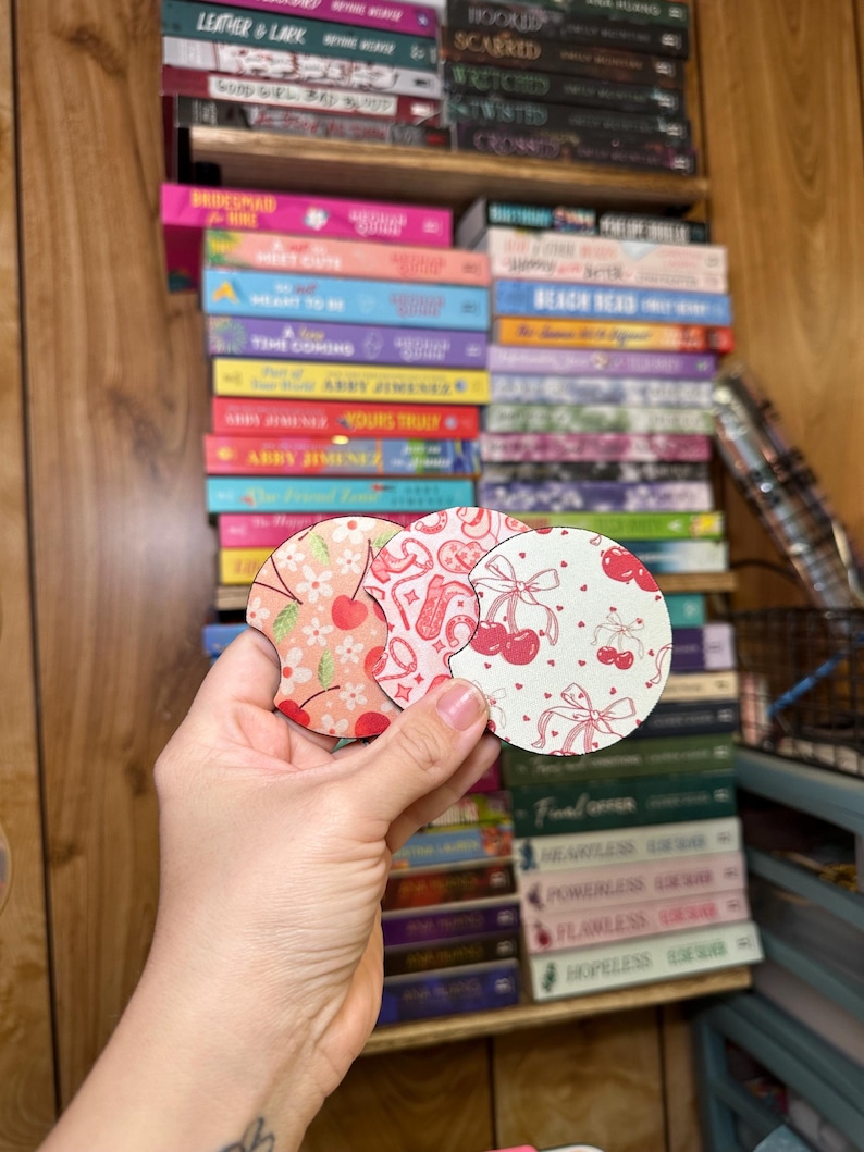 May include: Two round, pink and white coasters with a cherry and bow design. The coasters are made of wood and have a glossy finish.