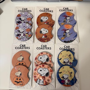 May include: Six sets of two circular car coasters in clear plastic packaging, each labeled CAR COASTERS. The coasters feature Snoopy and Woodstock in various autumn and Halloween-themed illustrations, including pumpkins, falling leaves, and witch hats.