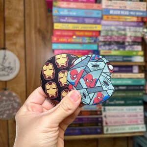 May include: Two round drink coasters featuring a superhero theme. One coaster has a blue, black, and red design with a repeated image of Spiderman. The other coaster has a red, yellow, and black design with a repeated image of Iron Man.