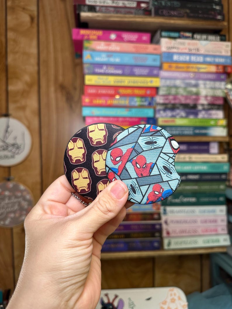 May include: Two round drink coasters featuring a superhero theme. One coaster has a blue, black, and red design with a repeated image of Spiderman. The other coaster has a red, yellow, and black design with a repeated image of Iron Man.