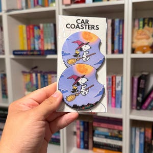 May include: Two round car coasters in clear packaging labeled CAR COASTERS. The coasters feature a graphic of Snoopy and Woodstock wearing witch hats and flying on a broomstick against a purple sky with a large orange full moon and clouds.