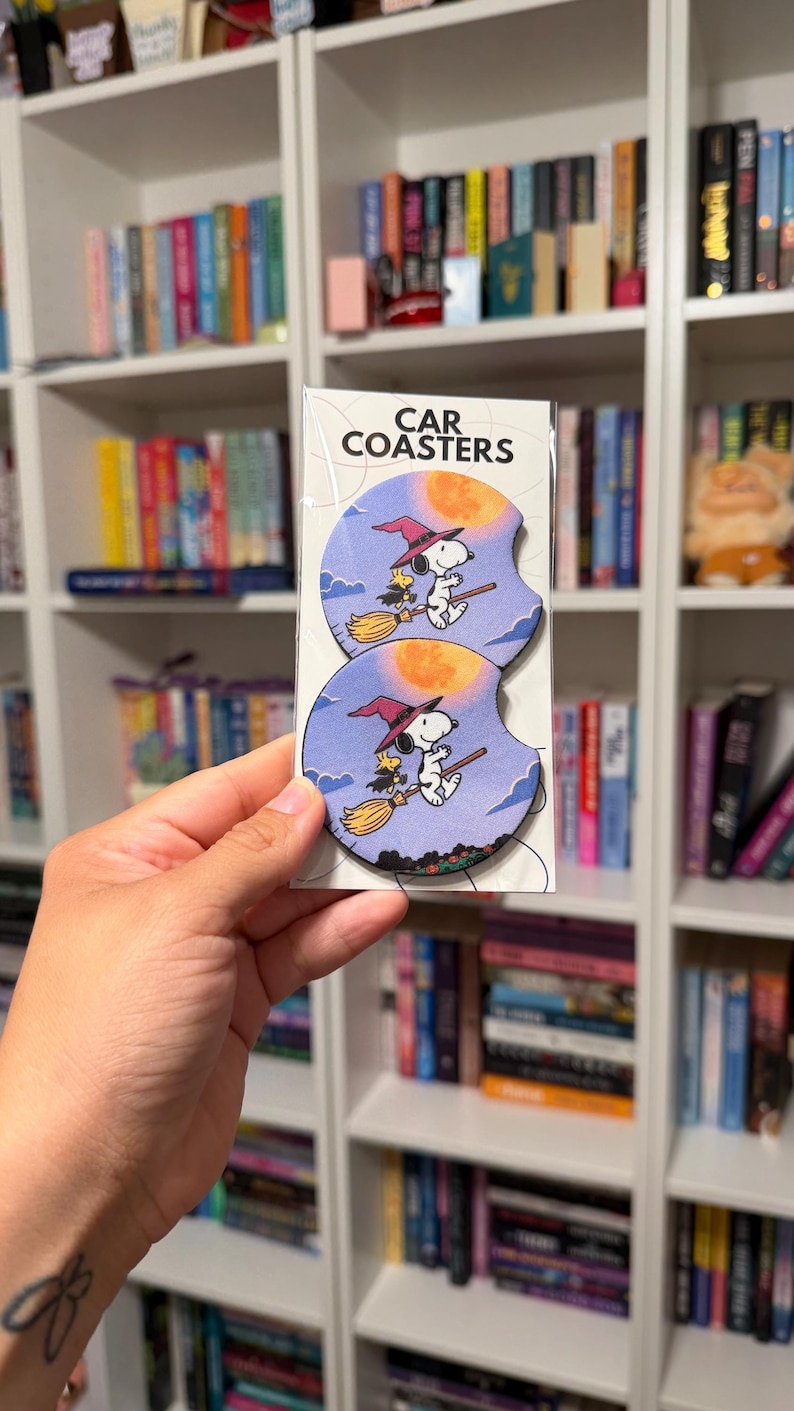May include: Two round car coasters in clear packaging labeled CAR COASTERS. The coasters feature a graphic of Snoopy and Woodstock wearing witch hats and flying on a broomstick against a purple sky with a large orange full moon and clouds.